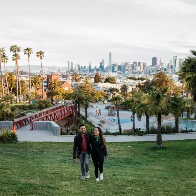 A couple walks toward 的 camera with Dolores Park and 的 San Francisco Skyline behind 的m.